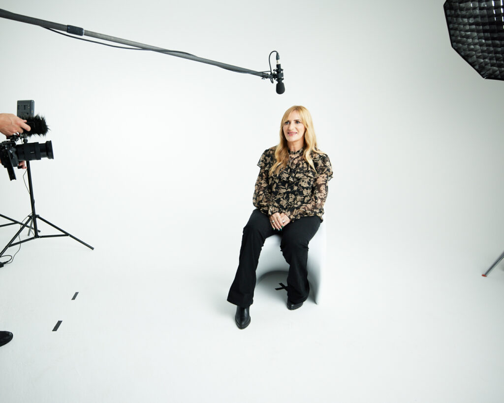 Bernadette sits in a chair on a film set. A large mic hangs above her. 